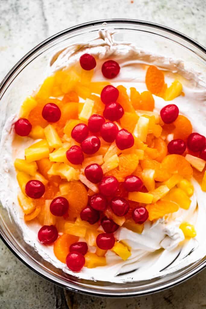 overhead shot of whipped topping in a bowl with mandarin oranges, maraschino cherries, and pineapple tidbits arranged on top.