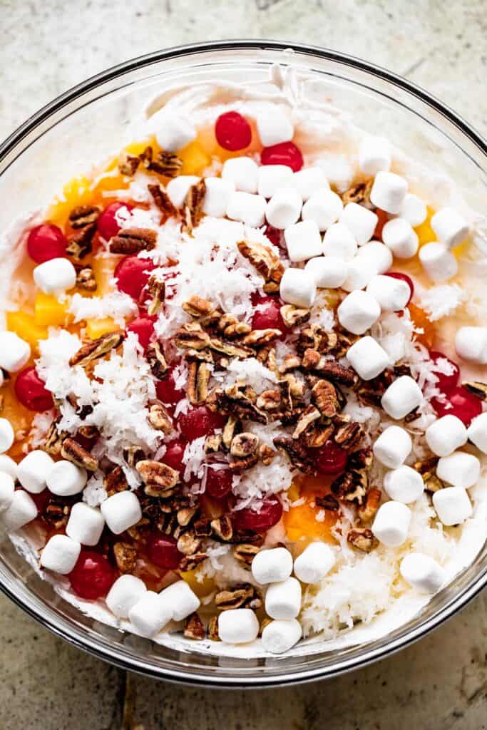 overhead shot of a bowl with whipped topping andchopped pecans, mini marshmallows, maraschino cherries, and mandarin oranges set on top.