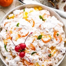 overhead shot of Ambrosia Salad served in a white dish and topped with three maraschino cherries and mint leaves. A bowl of chopped pecans are set above the salad, with two spoons next to it, and a mandarin orange.