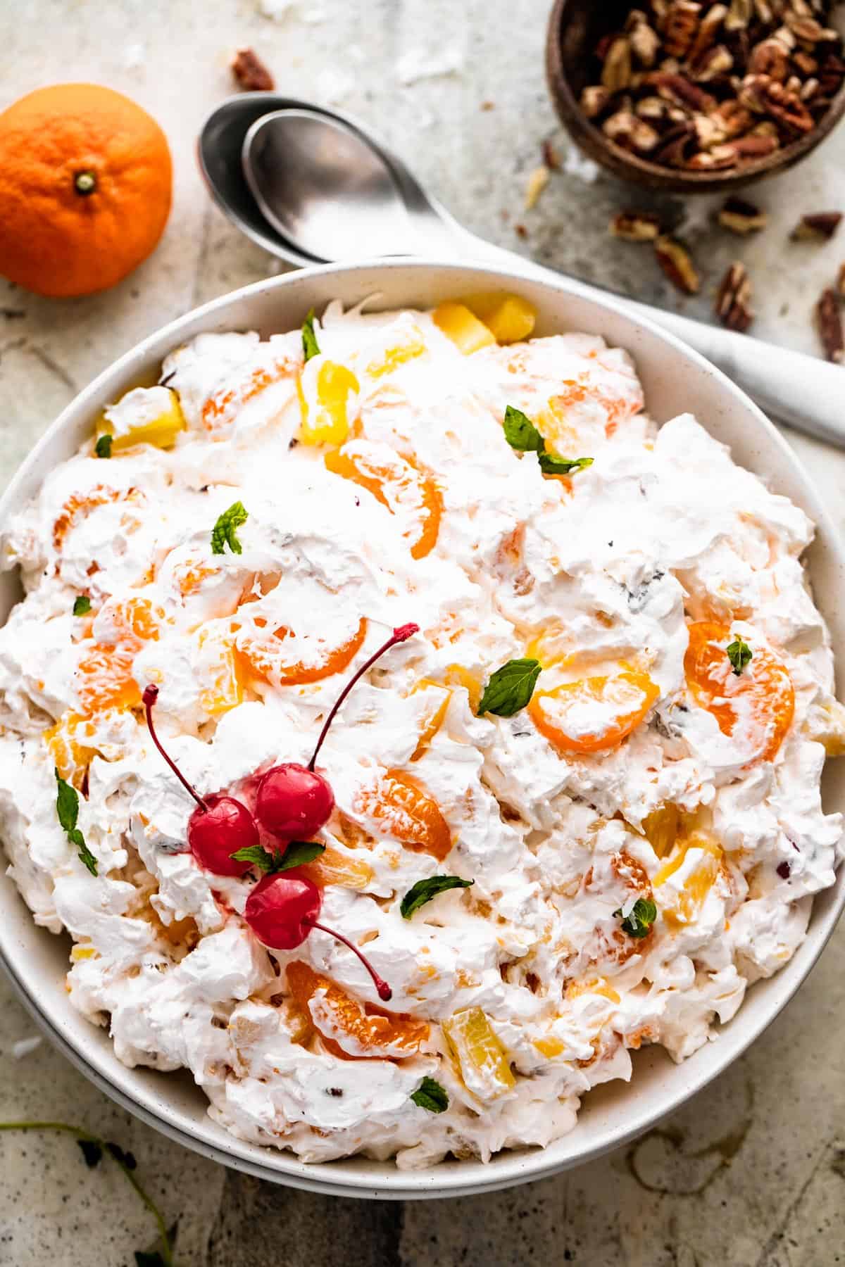 overhead shot of Ambrosia Salad served in a white dish and topped with three maraschino cherries and mint leaves. A bowl of chopped pecans are set above the salad, with two spoons next to it, and a mandarin orange.
