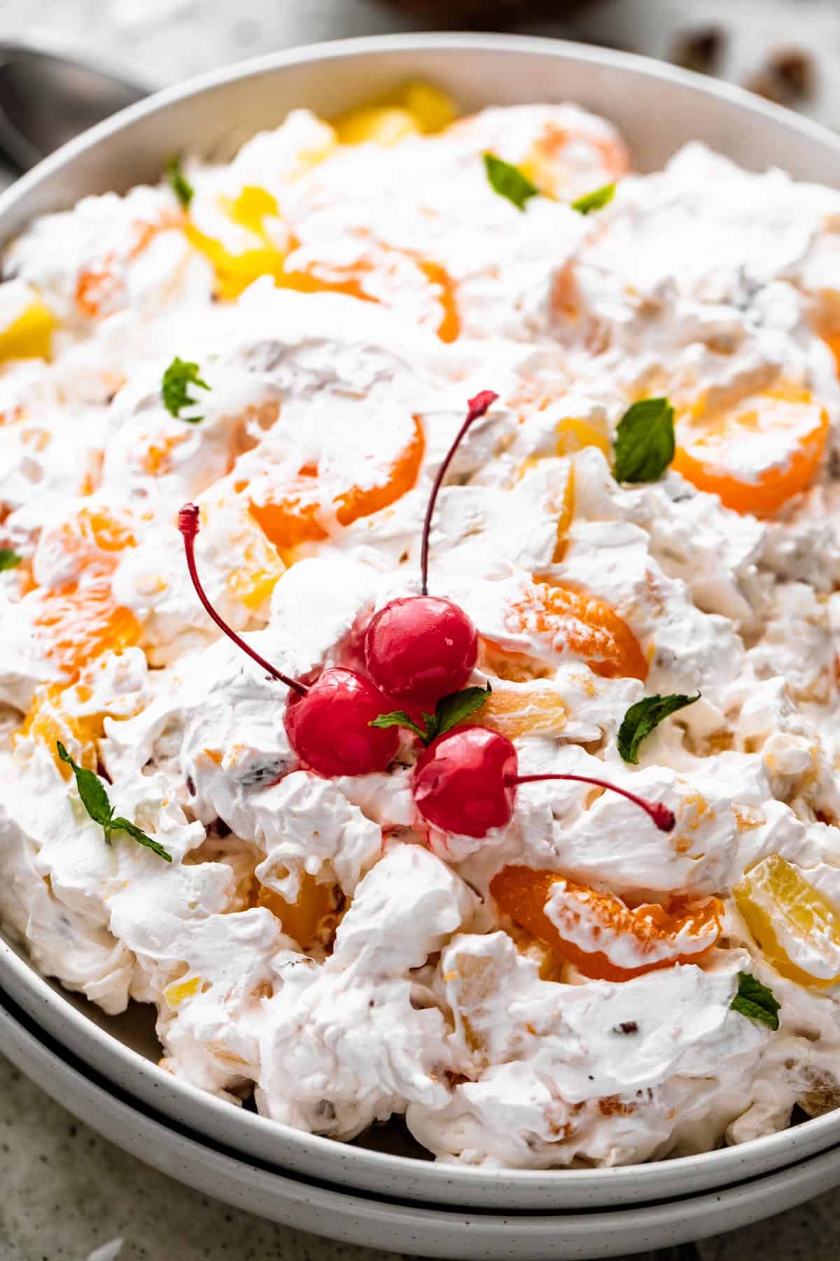 close up side shot of Ambrosia Salad served in a white dish and topped with three maraschino cherries and mint leaves.