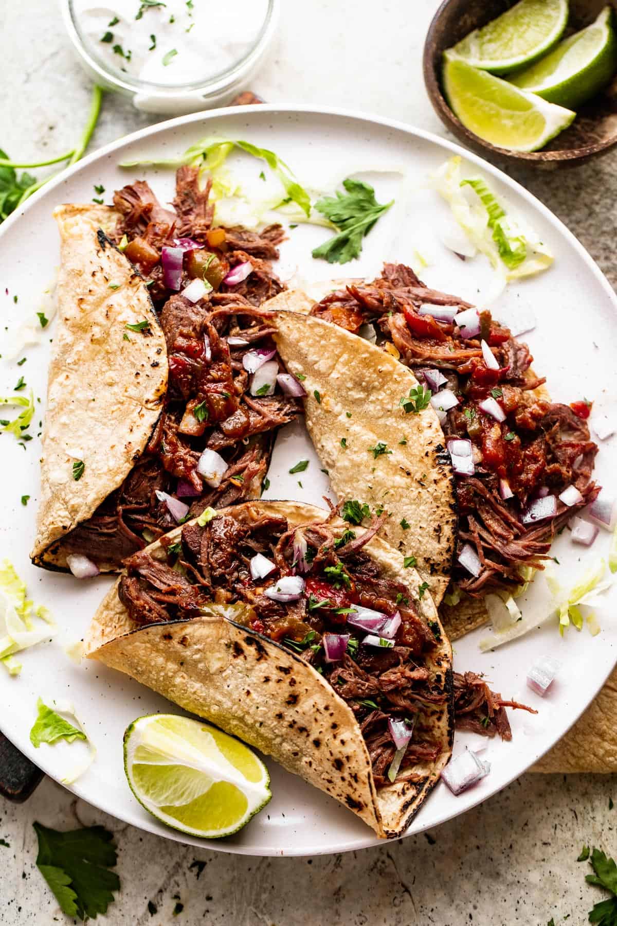 overhead shot of three Slow Cooker Beef Machaca tacos arranged on a round white plate with a small bowl of lime wedges placed next to the plate.