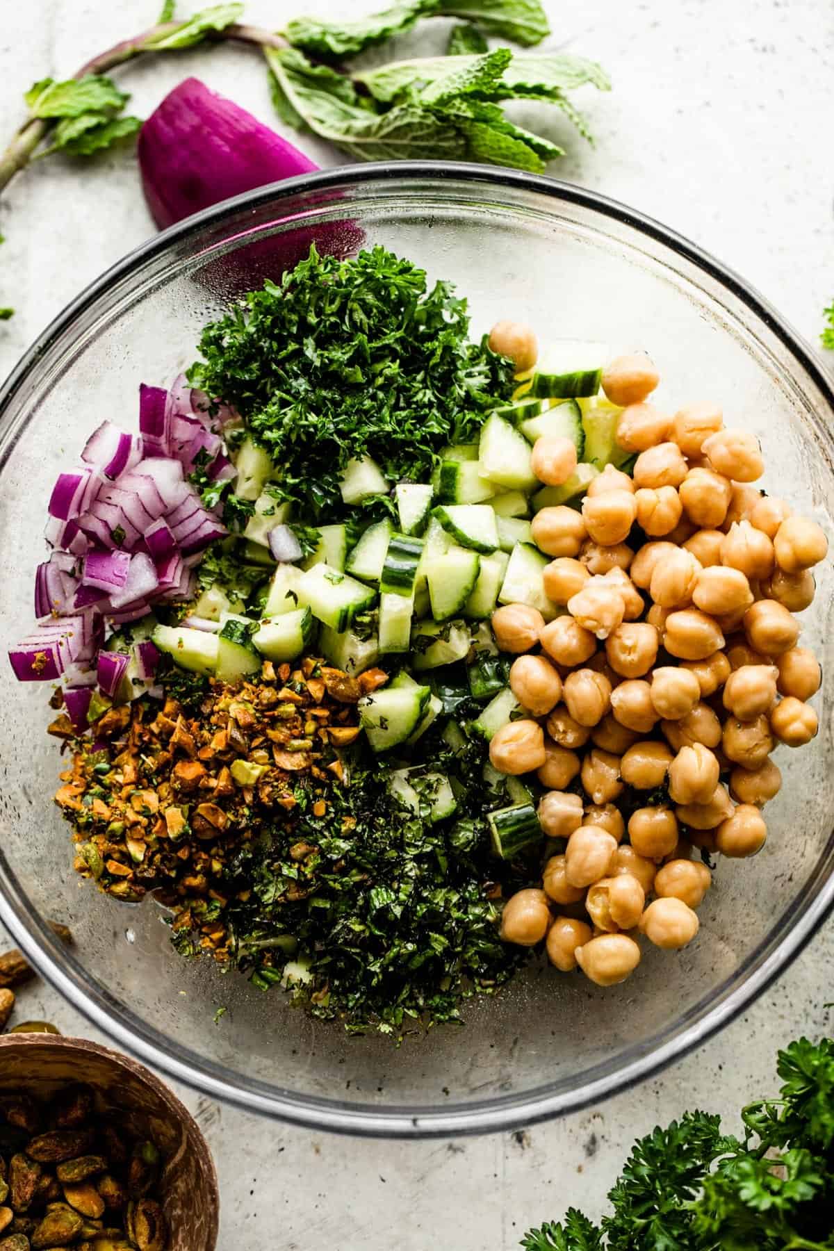 a glass bowl with chickpeas, mint, parsley, diced cucumbers, chopped red onions, and chopped pistachios.