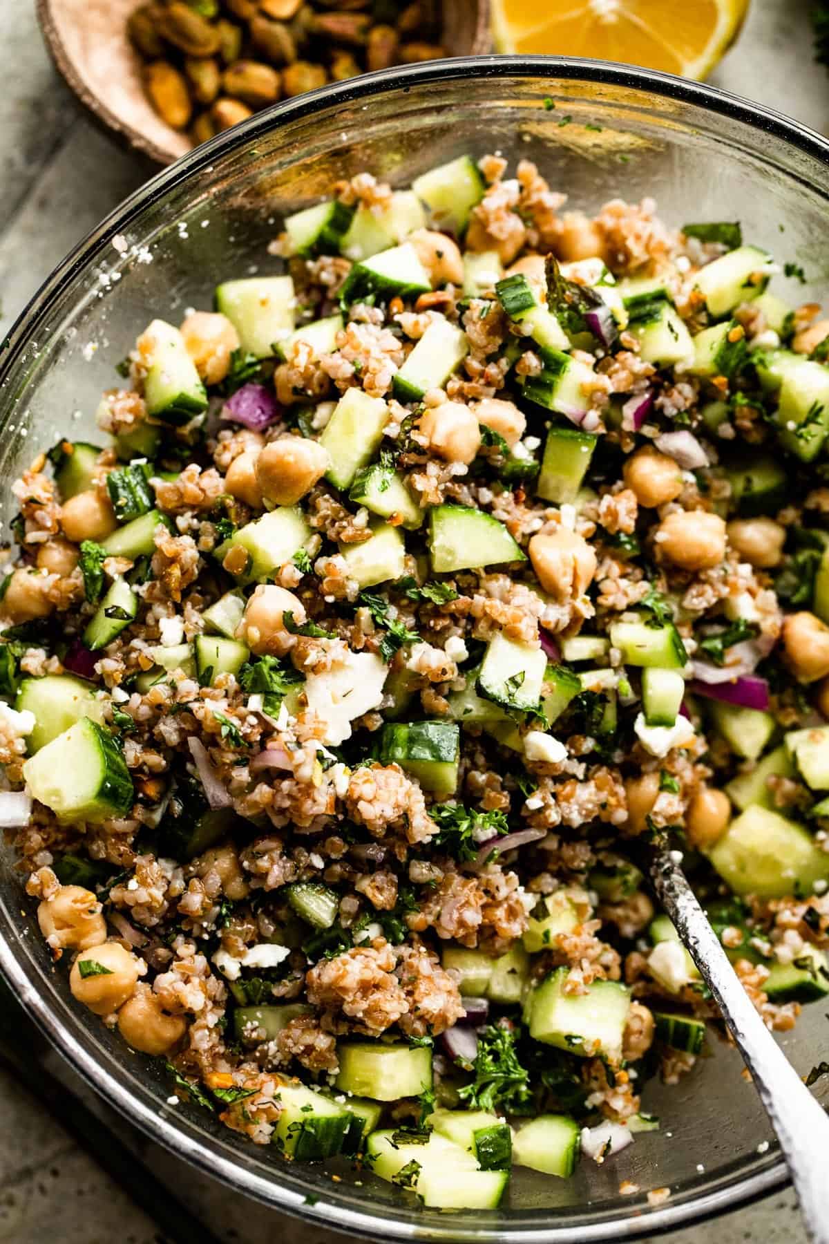 Up close shot of a glass mixing bowl with chickpeas, cucumbers, bulgur, and red onions.