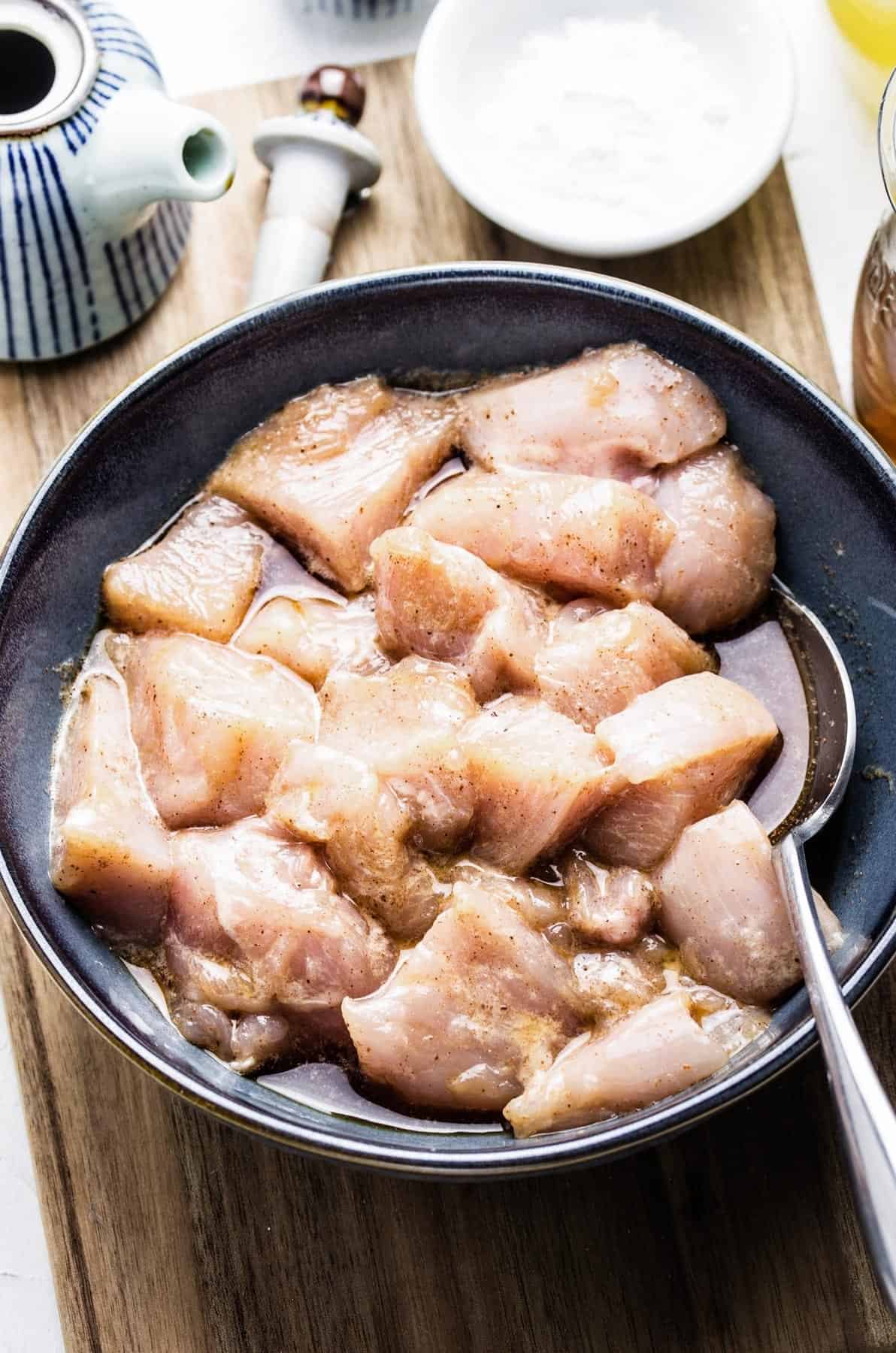 Cubes of raw chicken marinating in a bowl with a metal spoon inside.