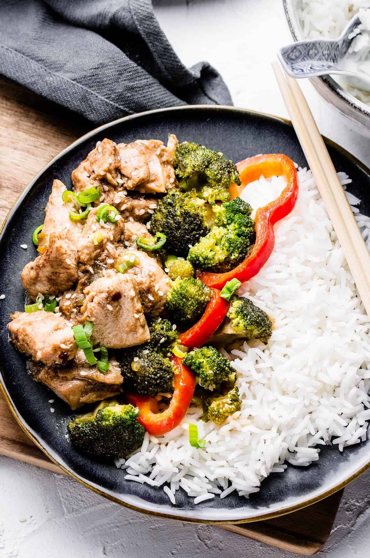 A plate of Chinese chicken and broccoli with white rice and a pair of chopsticks.