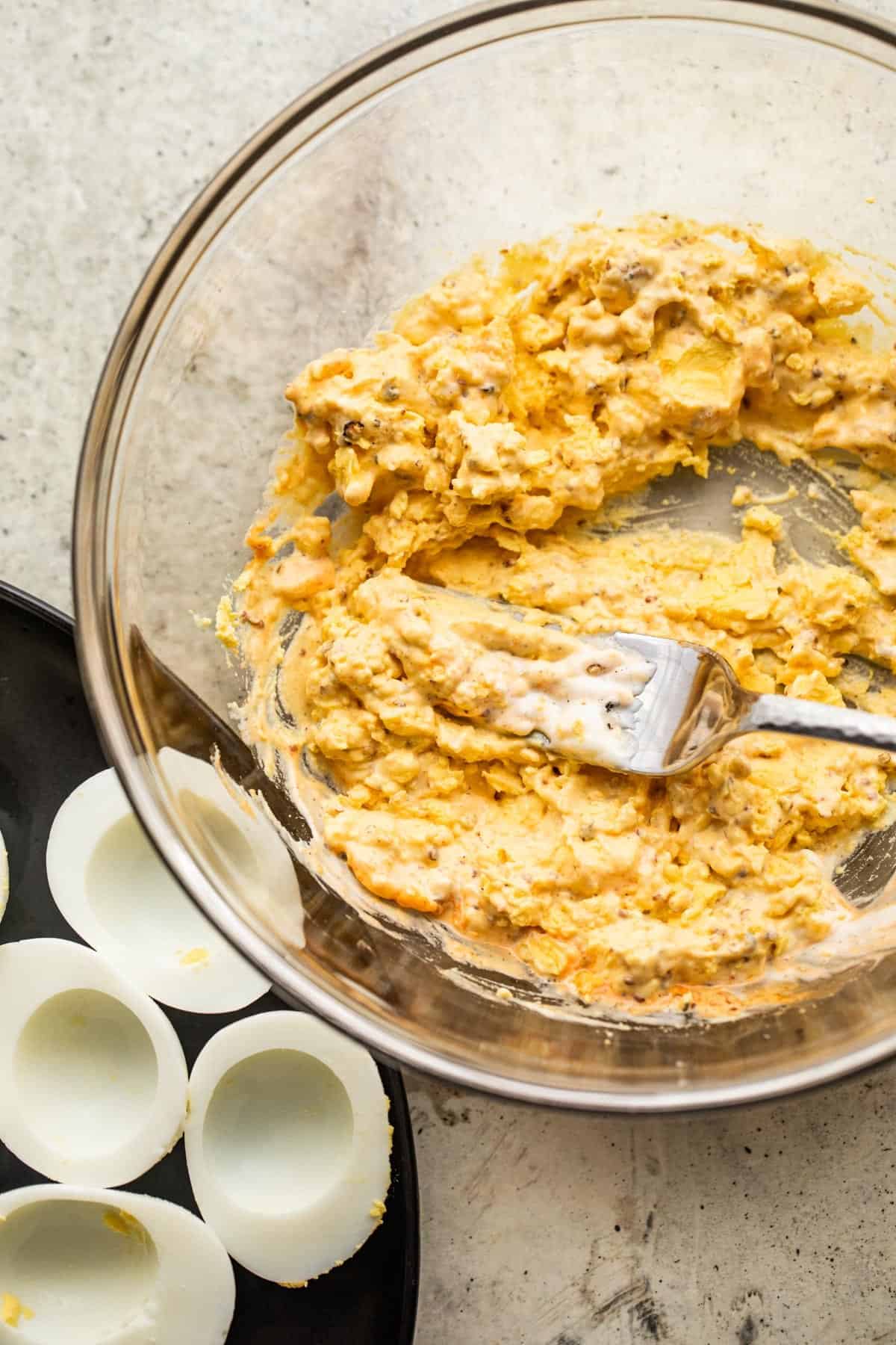 A fork is mashing egg yolks in a glass bowl, and a black plate with the egg whites is set on the left side of the bowl.