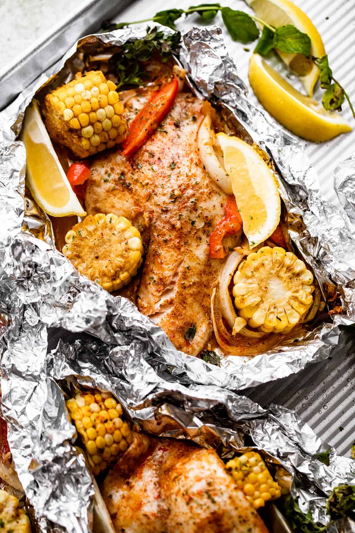 overhead shot of grilled tilapia arranged in foil packets with corn on the cob, lemon slices, and veggies arranged around the fish.