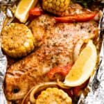 overhead shot of grilled tilapia filet set on a piece of foil and corn on the cob, lemon slices, and veggies arranged around the fish.