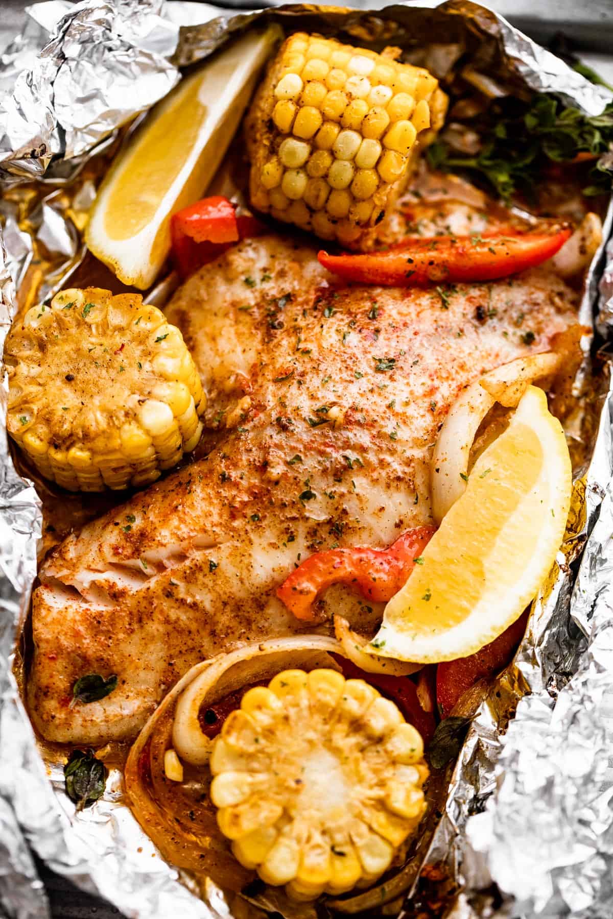 overhead shot of grilled tilapia filet set on a piece of foil and corn on the cob, lemon slices, and veggies arranged around the fish.