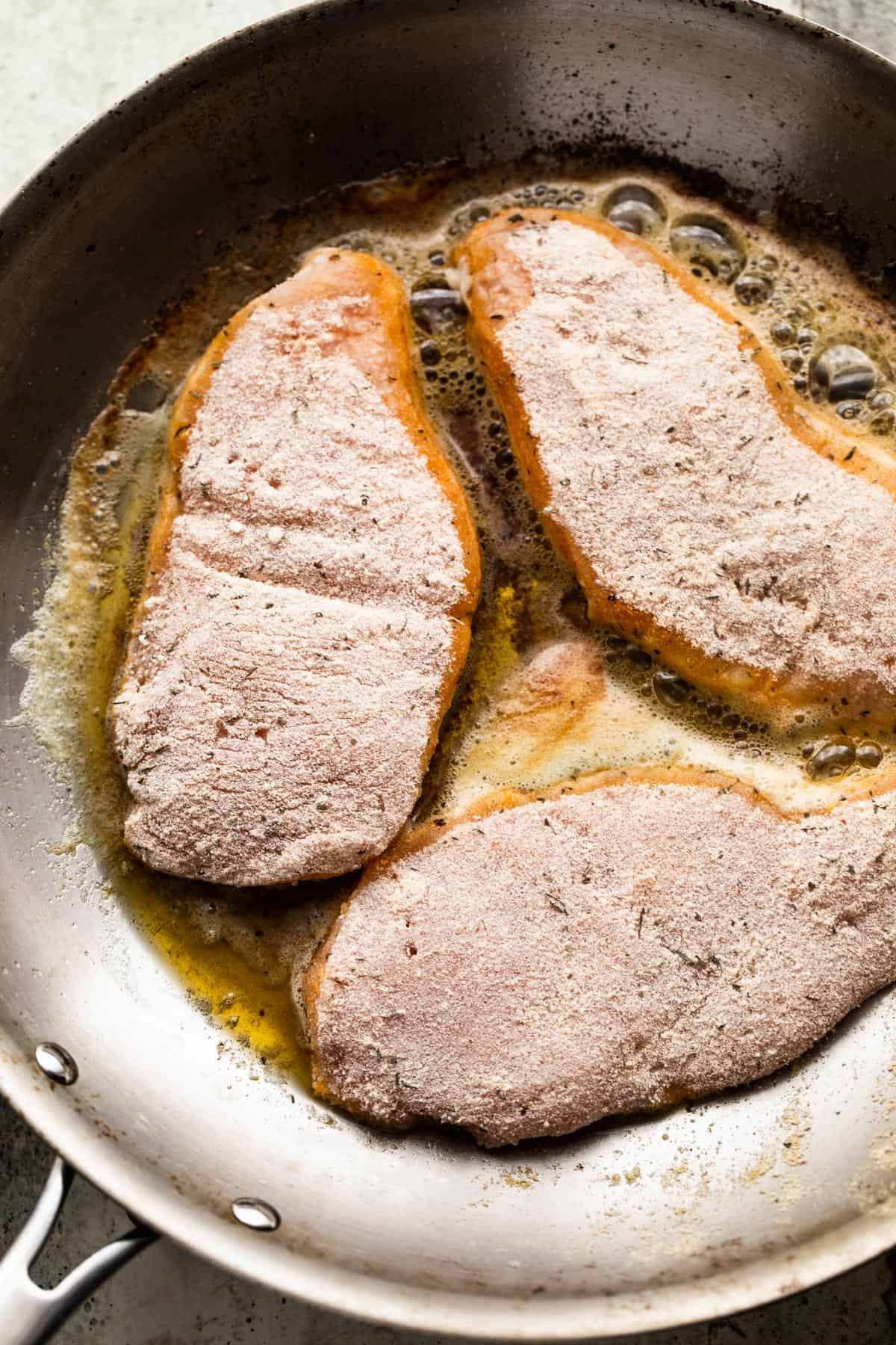 Three breaded chicken breasts frying in a skillet.