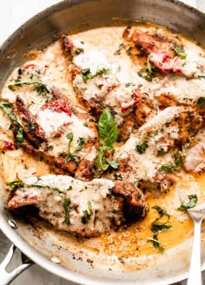 a skillet with five chicken cutlets in a creamy sauce with sundried tomatoes, and garnished with chopped basil.