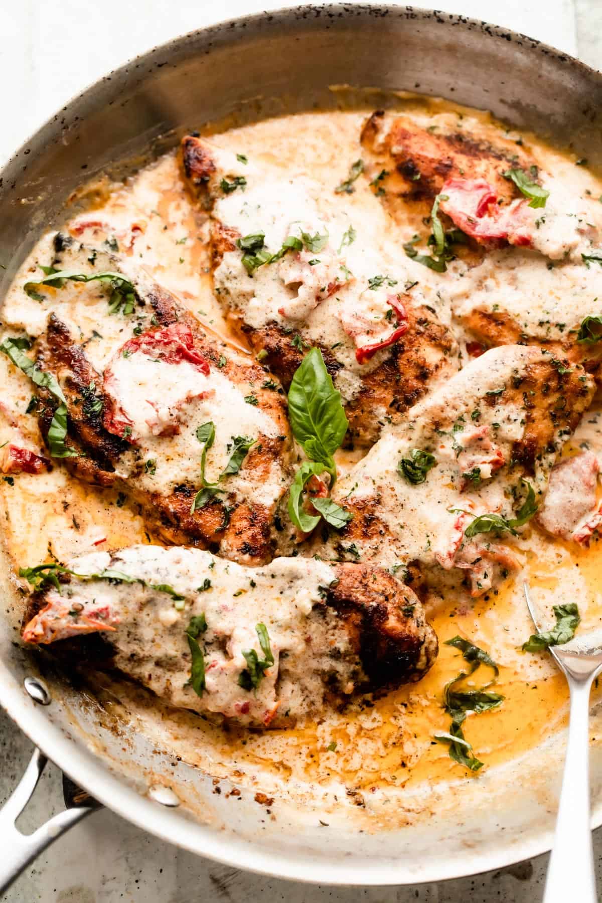 a skillet with five chicken cutlets in a creamy sauce with sundried tomatoes, and garnished with chopped basil.