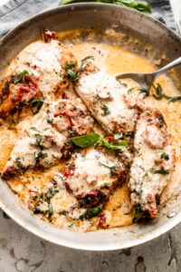 a skillet with five chicken cutlets in a creamy sauce with sundried tomatoes, and garnished with chopped basil.