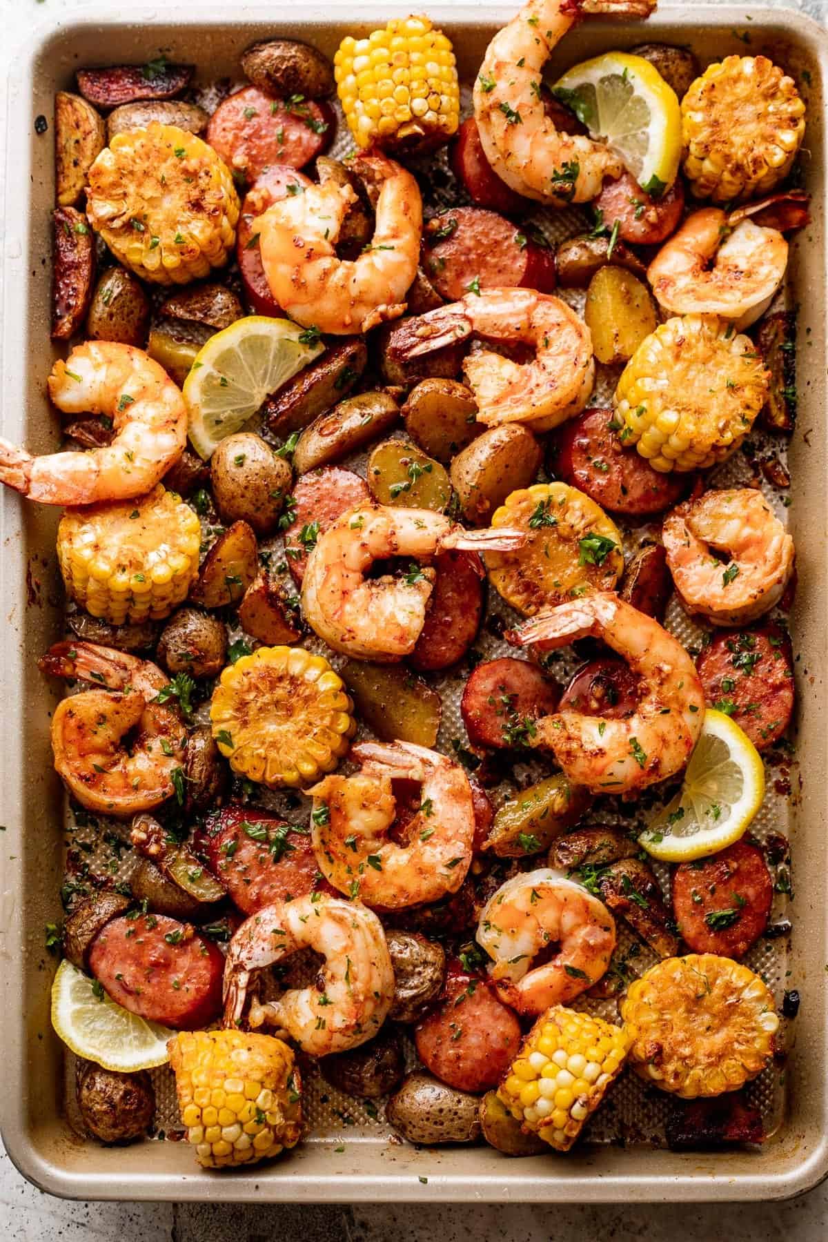 overhead shot of shrimp, potatoes, sausages, and corn arranged on a sheet pan and garnished with a few lemon slices.