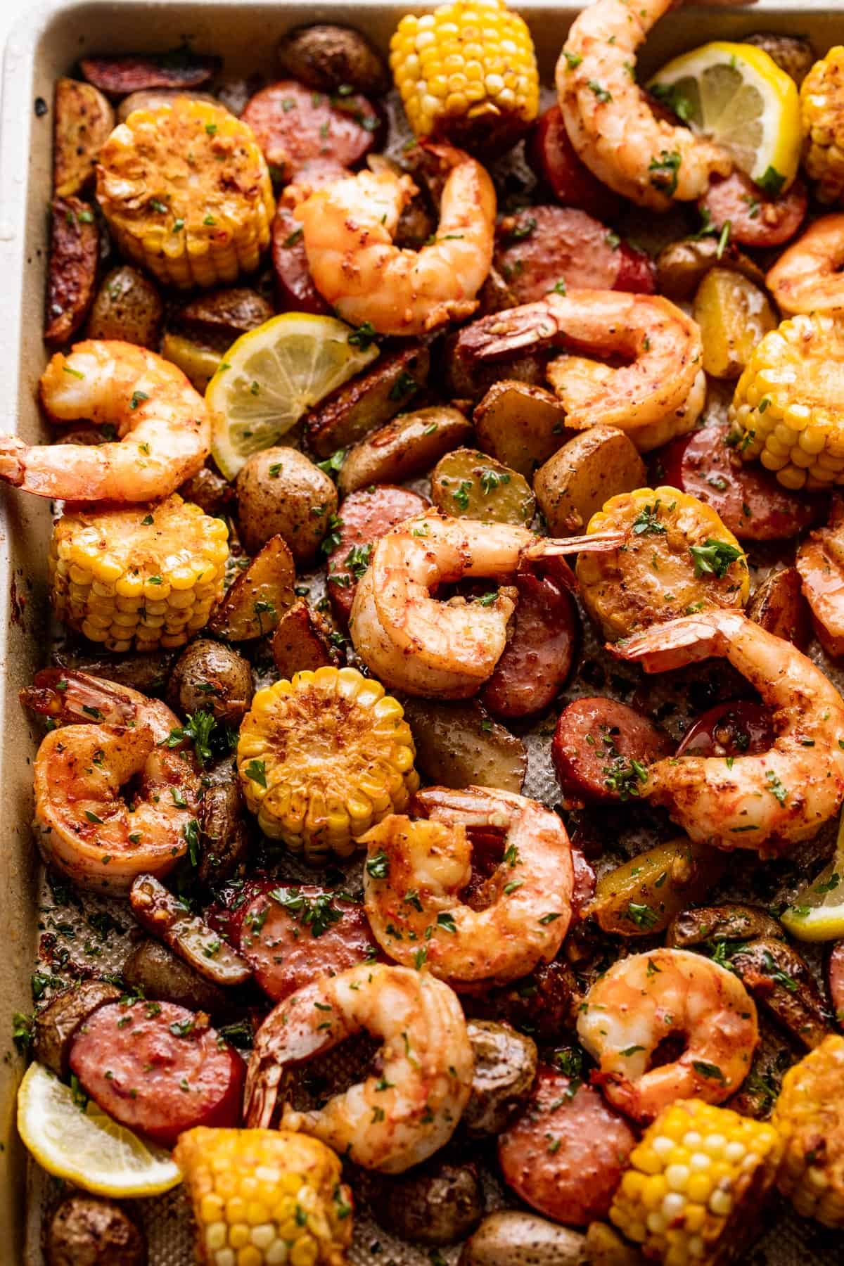 close up shot of shrimp, potatoes, sausages, and corn arranged on a sheet pan and garnished with a few lemon slices.
