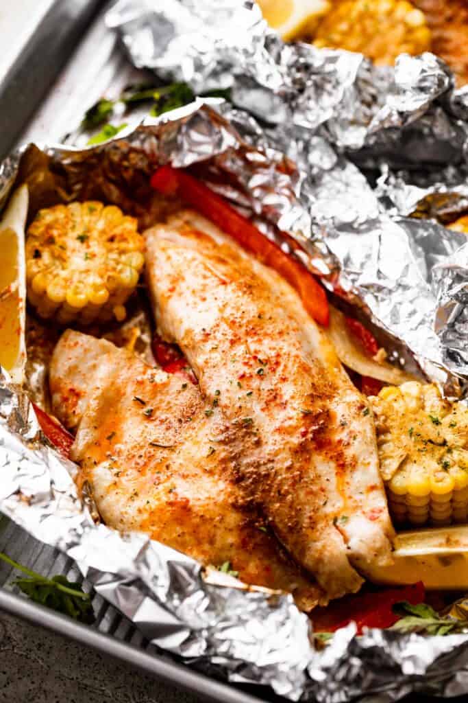 raw grilled tilapia filet set on a piece of foil and corn on the cob, lemon slices, and veggies arranged around the fish.