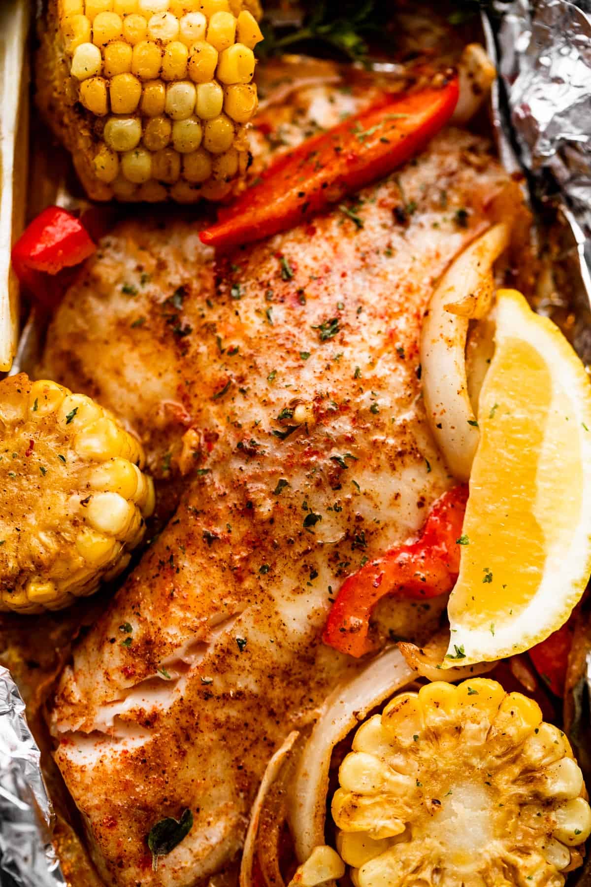 overhead shot of grilled tilapia filet set on a piece of foil and corn on the cob, lemon slices, and veggies arranged around the fish.