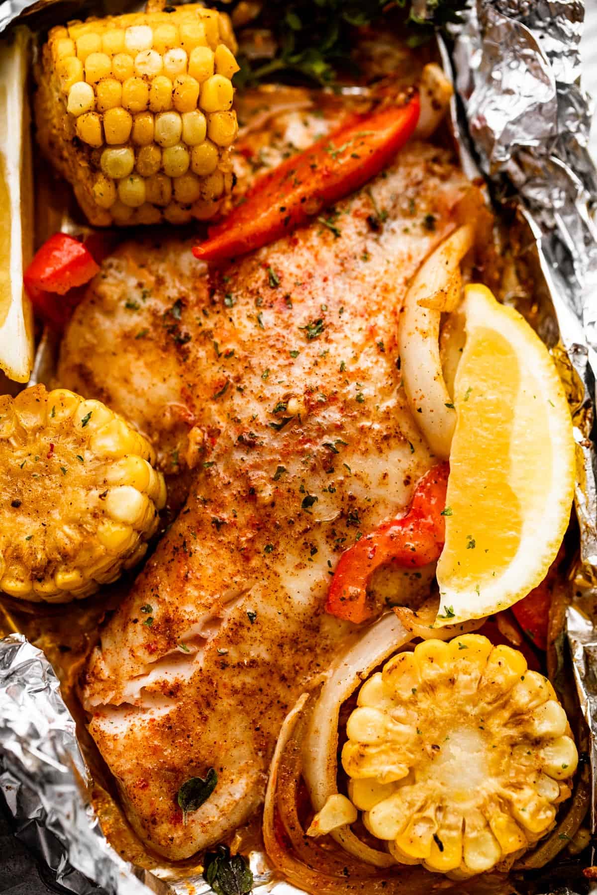 overhead shot of grilled tilapia filet set on a piece of foil and corn on the cob, lemon slices, and veggies arranged around the fish.