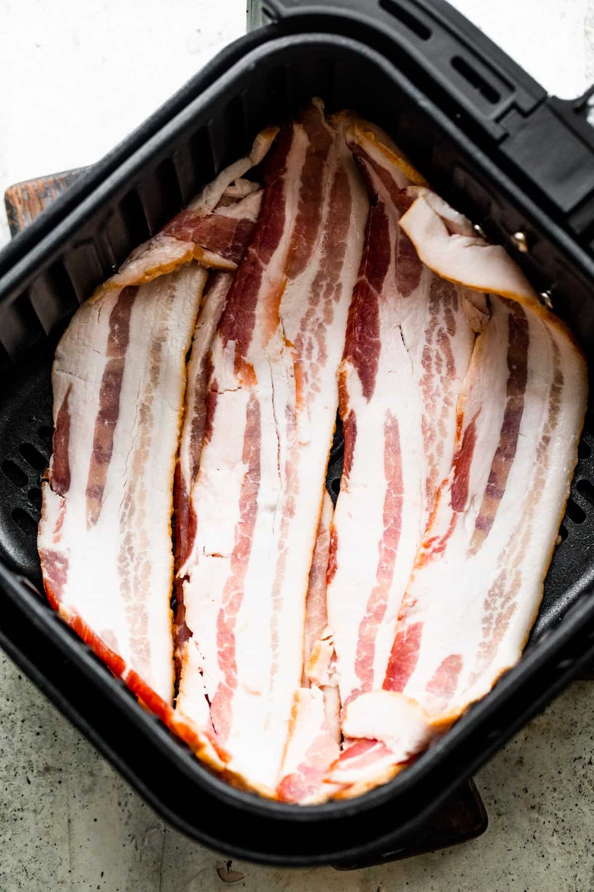raw bacon strips arranged inside of an air fryer basket.