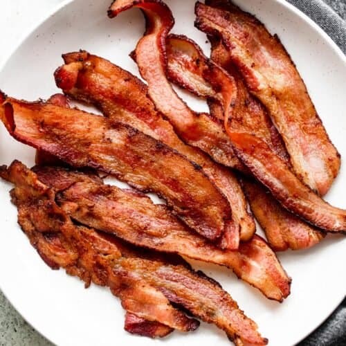 strips of air fryer bacon arranged on a white plate, with a gray tea towel placed underneath the plate.