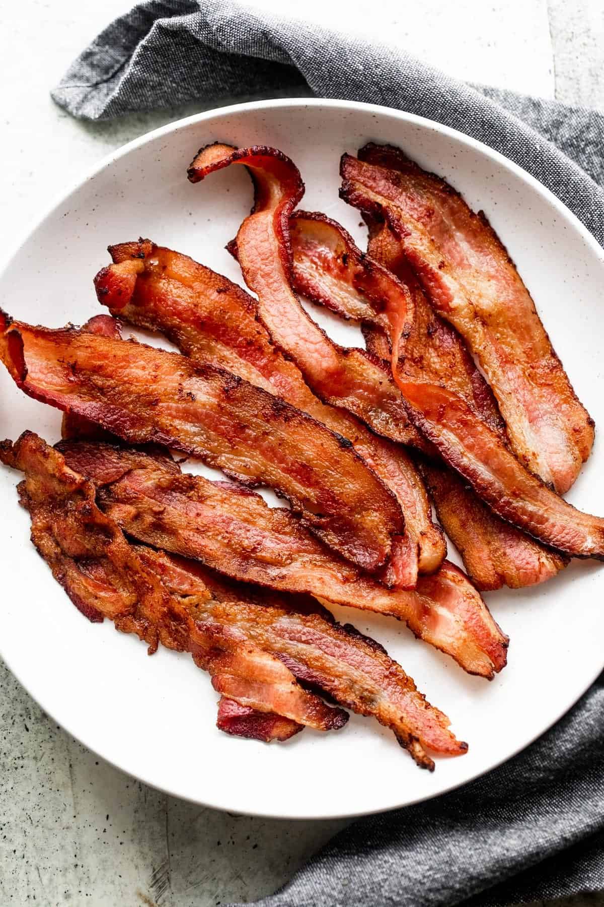 strips of air fryer bacon arranged on a white plate, with a gray tea towel placed underneath the plate.