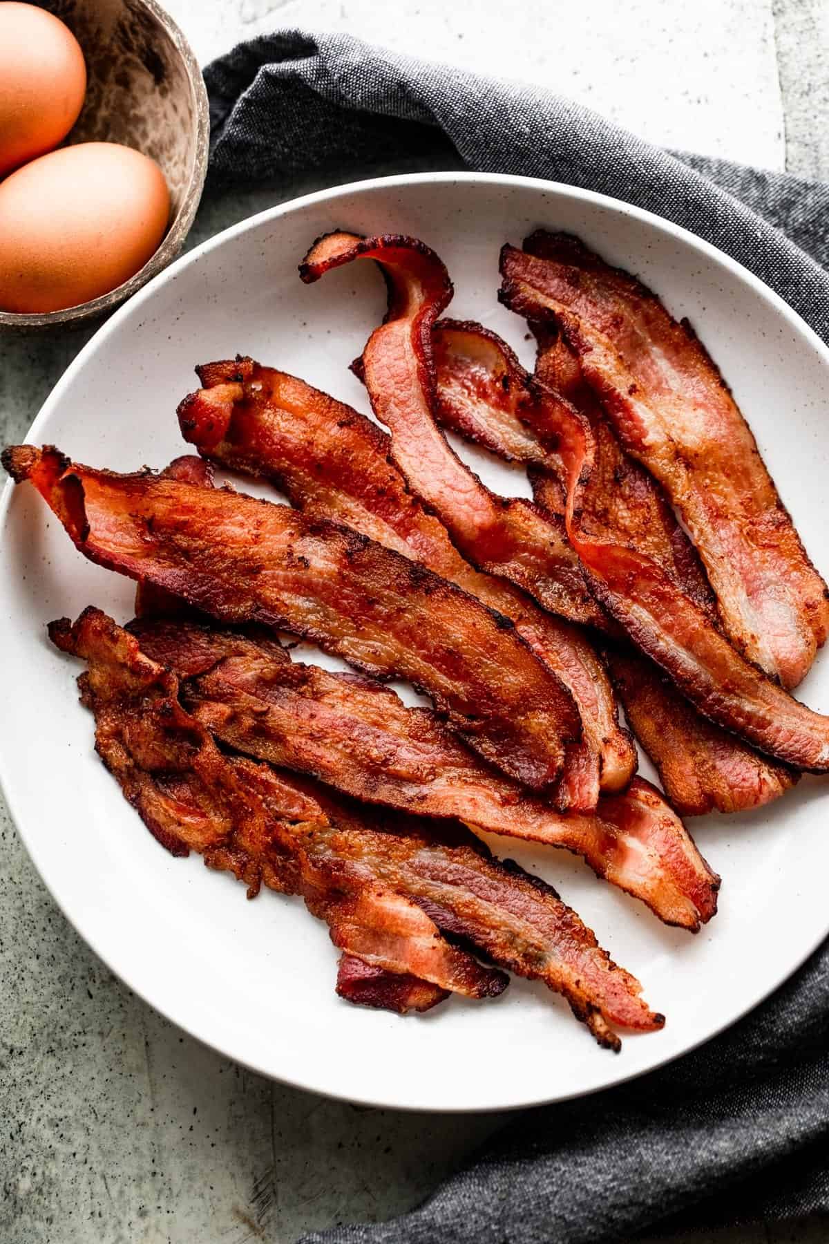 strips of air fried bacon arranged on a white serving plate that is set on a gray tea towel, and two eggs in a bowl placed to the left of the plate.
