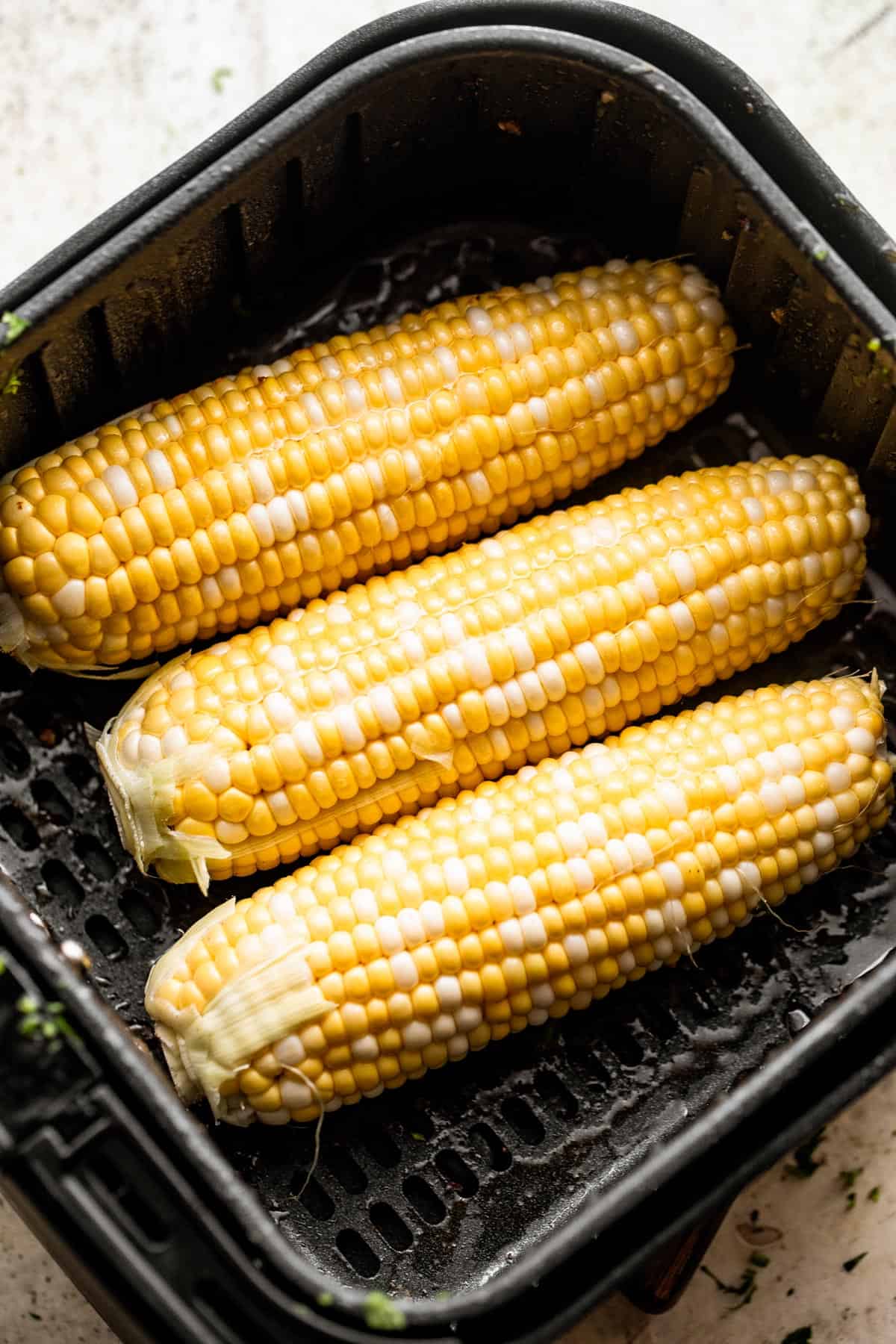 Black air fryer basket with three ears of raw corn arranged inside it.