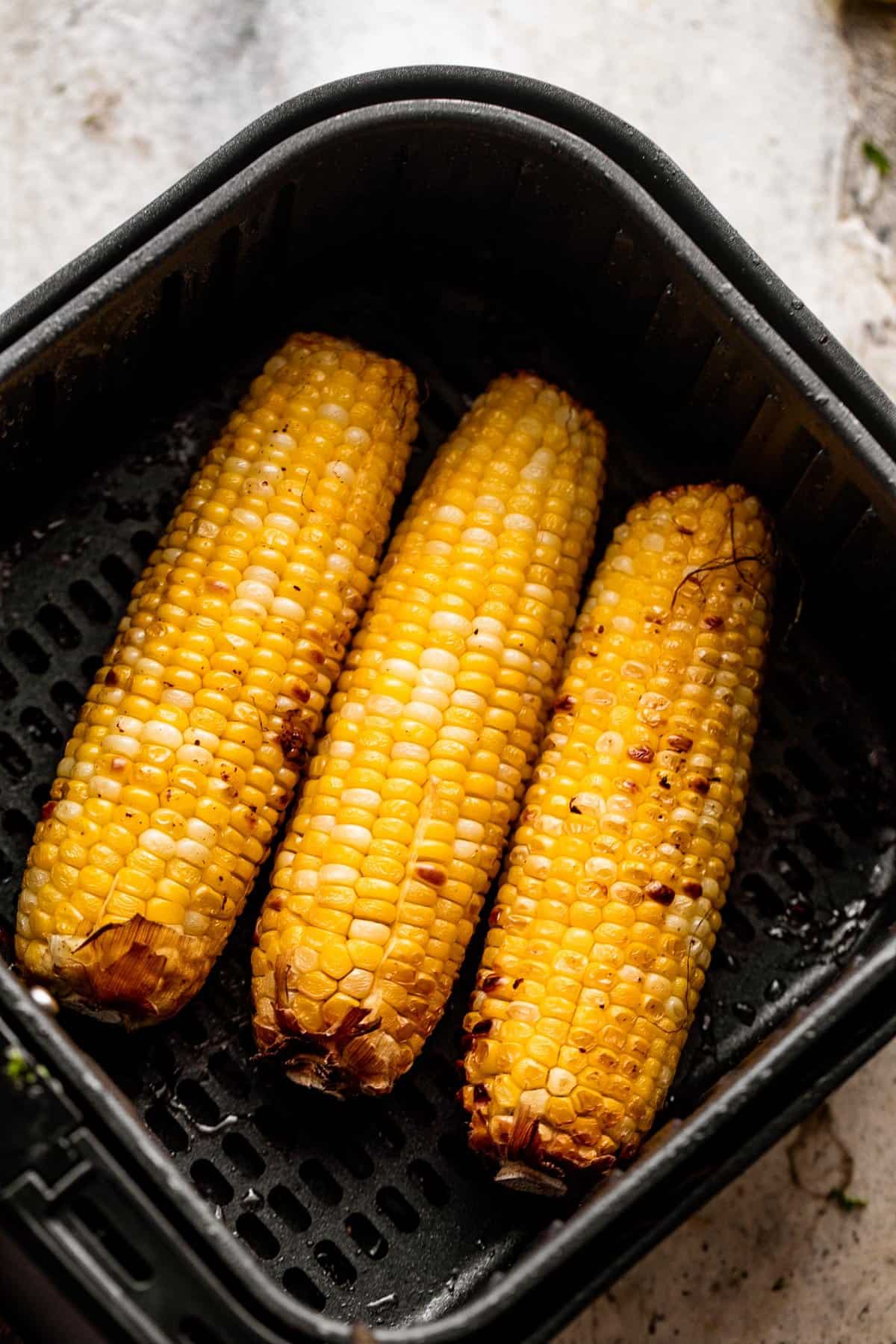 overhead shot of a black air fryer basket with three ears of corn arranged inside it.