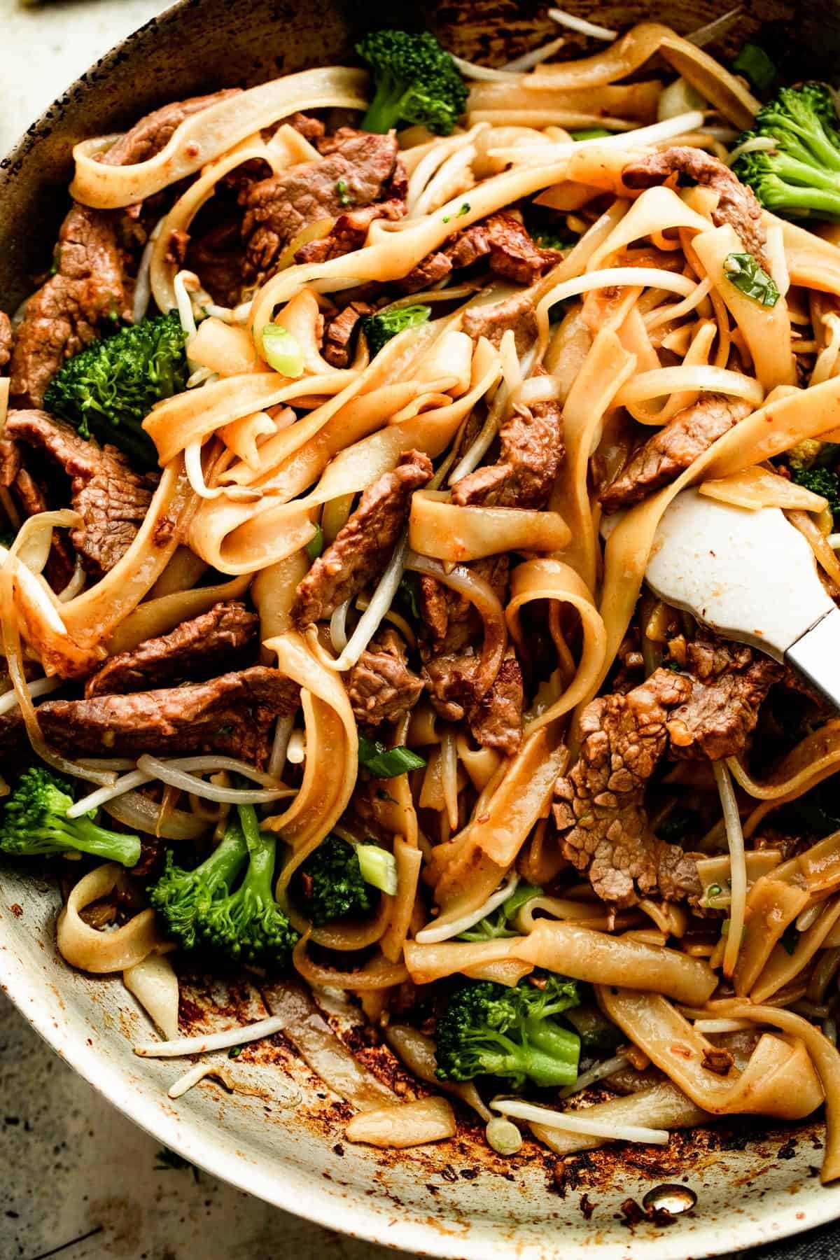 overhead shot of beef chow fun cooking in a skillet, with tongs resting in the middle.