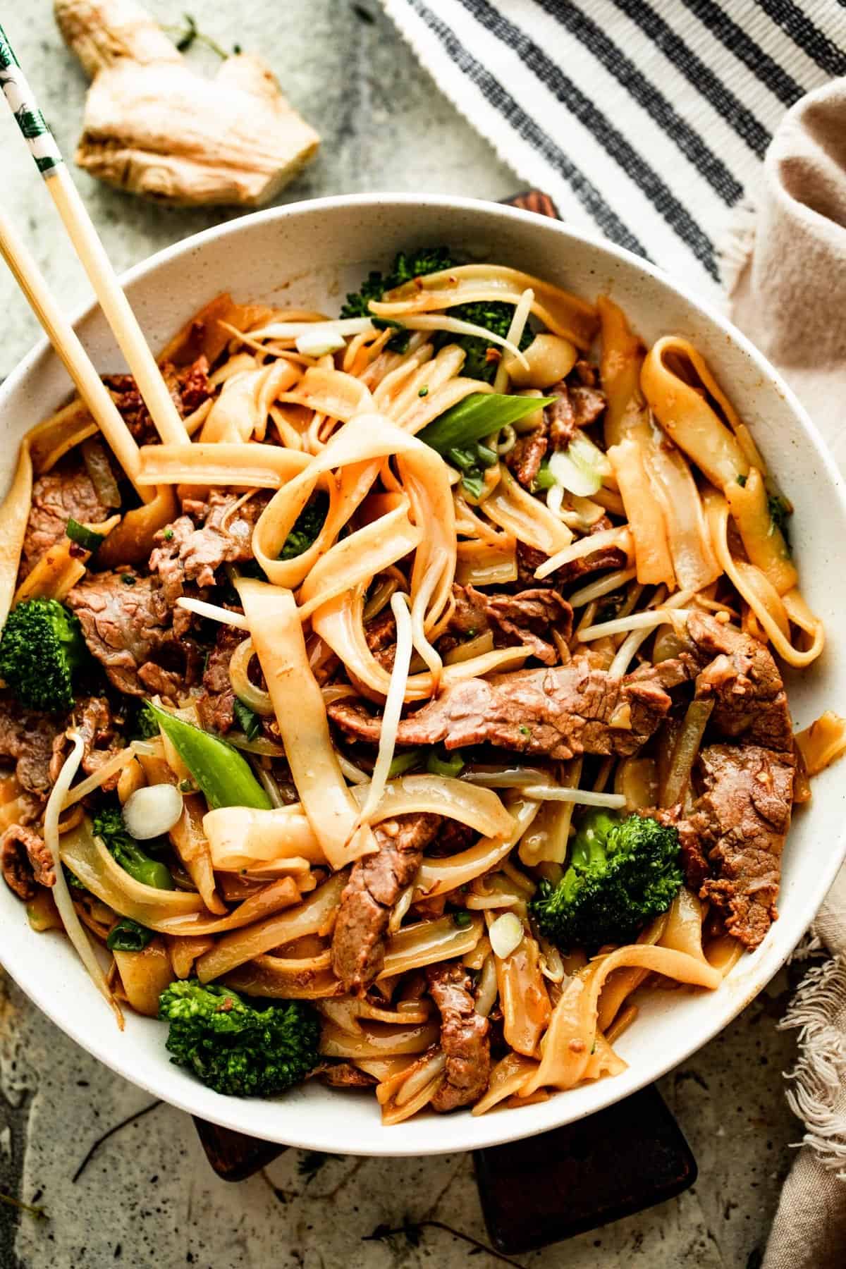 overhead shot of a white bowl with beef chow fun and two chopsticks resting on the left side of the bowl.