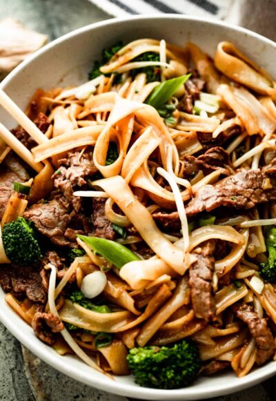 up close overhead shot of a white bowl with beef chow fun and two chopsticks resting on the left side of the bowl.