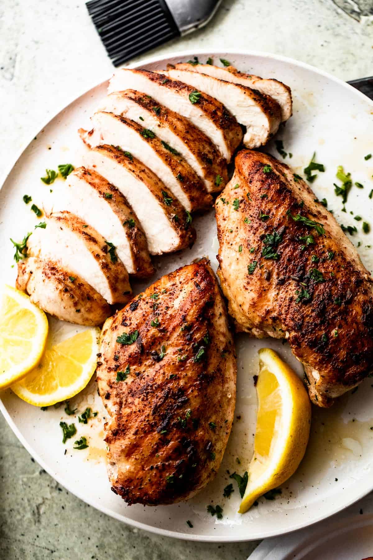 two whole grilled chicken breasts and one sliced grilled chicken breast served on a round white plate with lemon wedges.