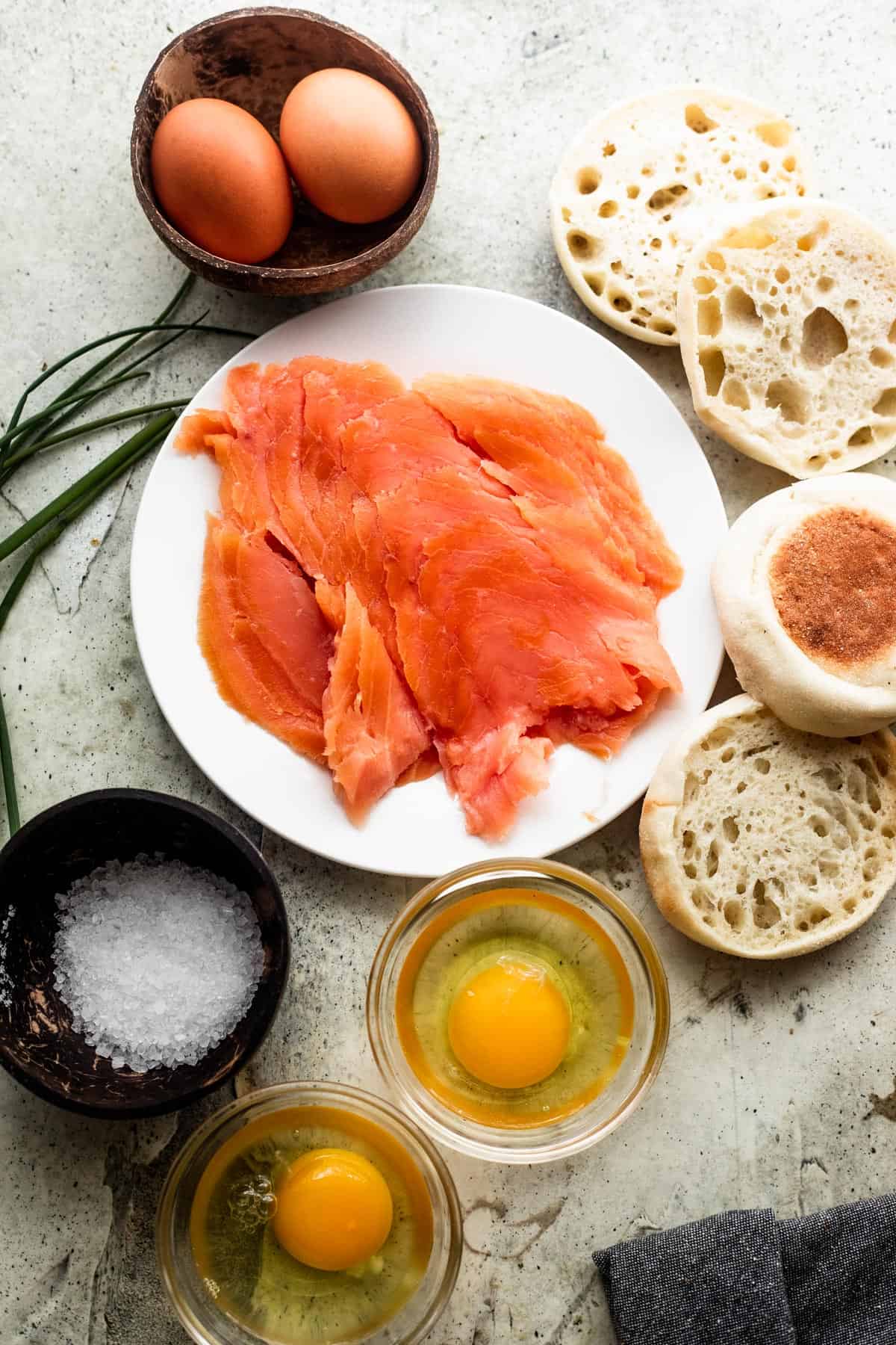 from left to right: two glass bowls with raw eggs inside, a small brown bowl with kosher salt, another small brown bowl with two brown eggs, and a row of english muffins split in half, with a plate of smoked salmon slices set in the center of everything.