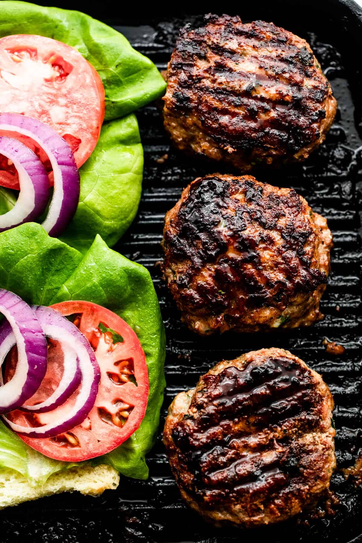 three turkey burgers on a black backdrop with lettuce, tomatoes, and red onion slices arranged alongside the burgers.