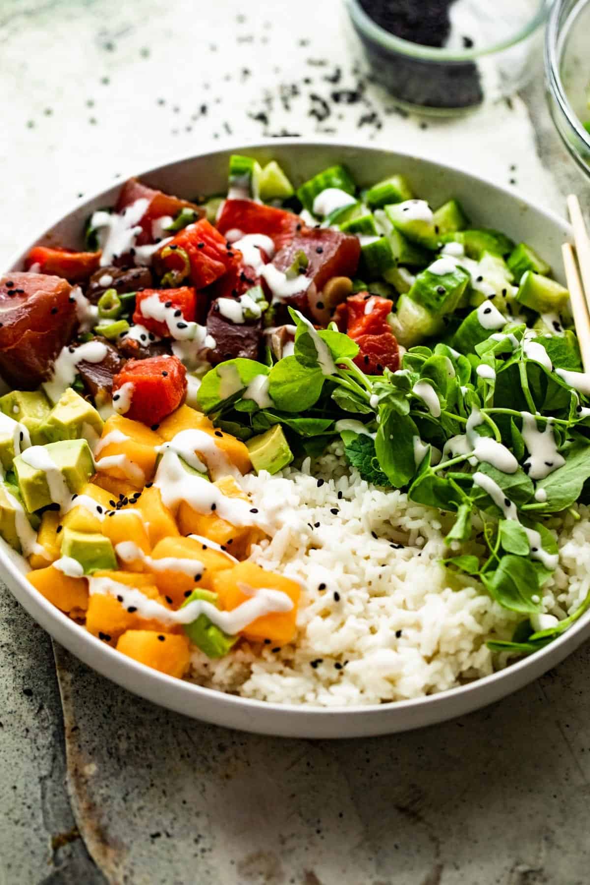 side shot of a poke bowl with rice, diced mango, diced tuna, diced salmon, diced cucumbers, diced avocados, and greens.