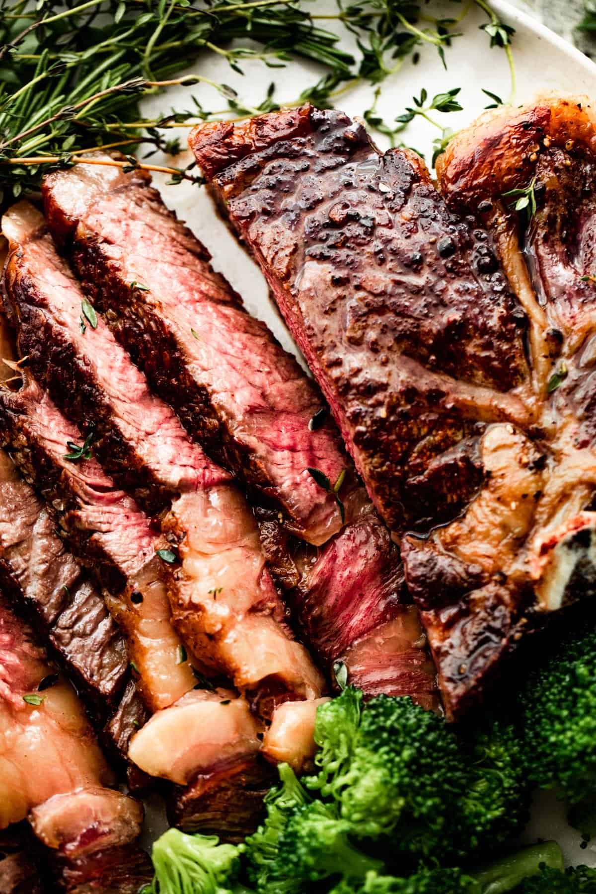 close up shot of a sliced ribeye steak on a white dinner plate with broccoli and greens arranged around the steak.