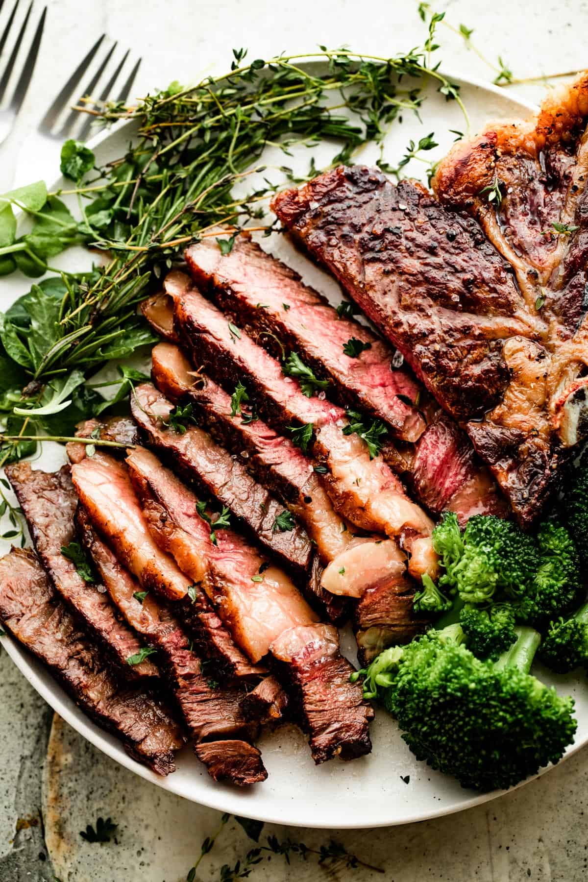 overhead shot of sliced ribeye steak on a white dinner plate with broccoli and greens arranged around the steak.