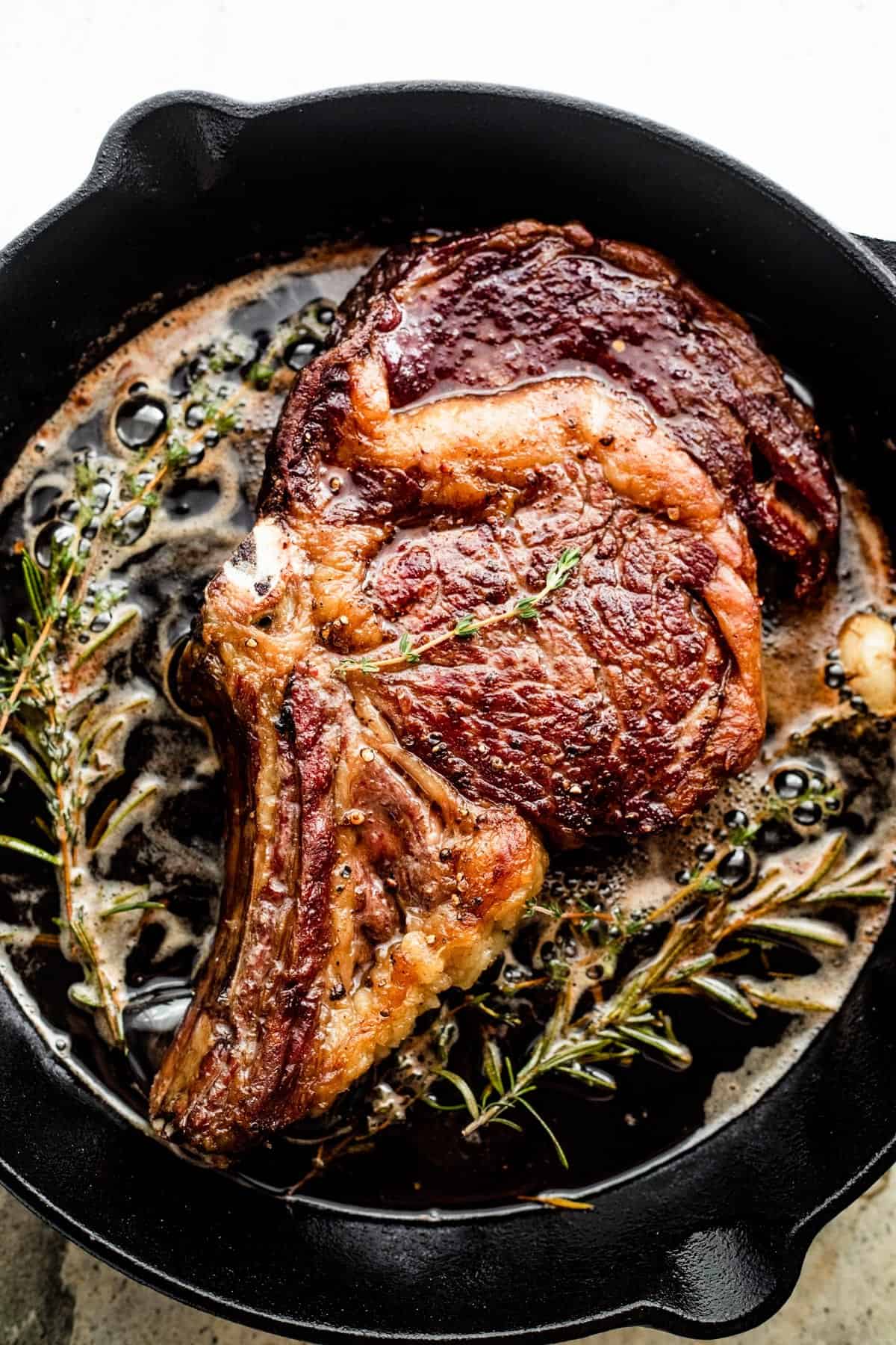 cooking a ribeye steak in a black cast iron skillet, with butter and herbs around the steak.
