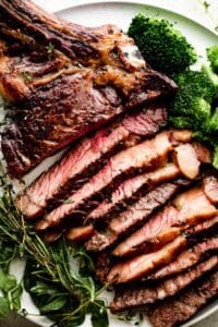 sliced ribeye steak on a white dinner plate with broccoli and greens arranged around the steak.