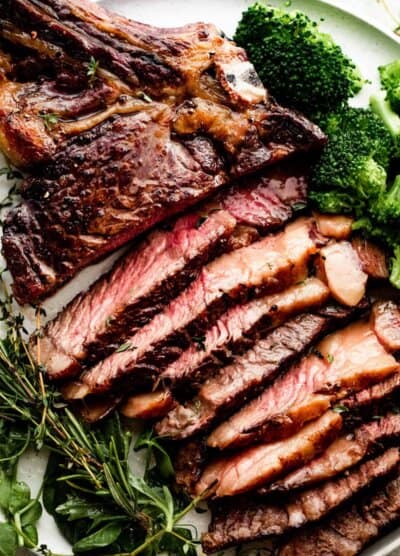 sliced ribeye steak on a white dinner plate with broccoli and greens arranged around the steak.