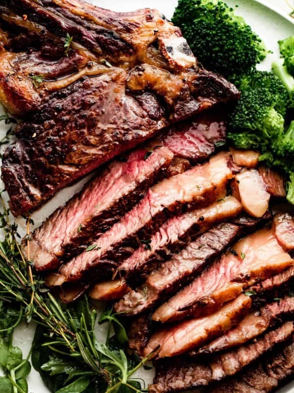 sliced ribeye steak on a white dinner plate with broccoli and greens arranged around the steak.