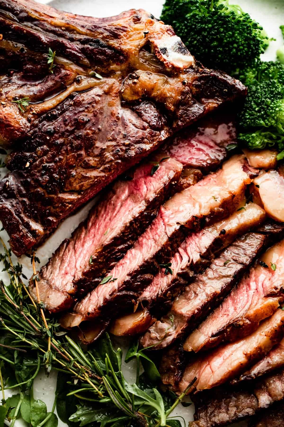 close up shot of a sliced reverse seared ribeye steak on a white dinner plate with broccoli and greens arranged around the steak.