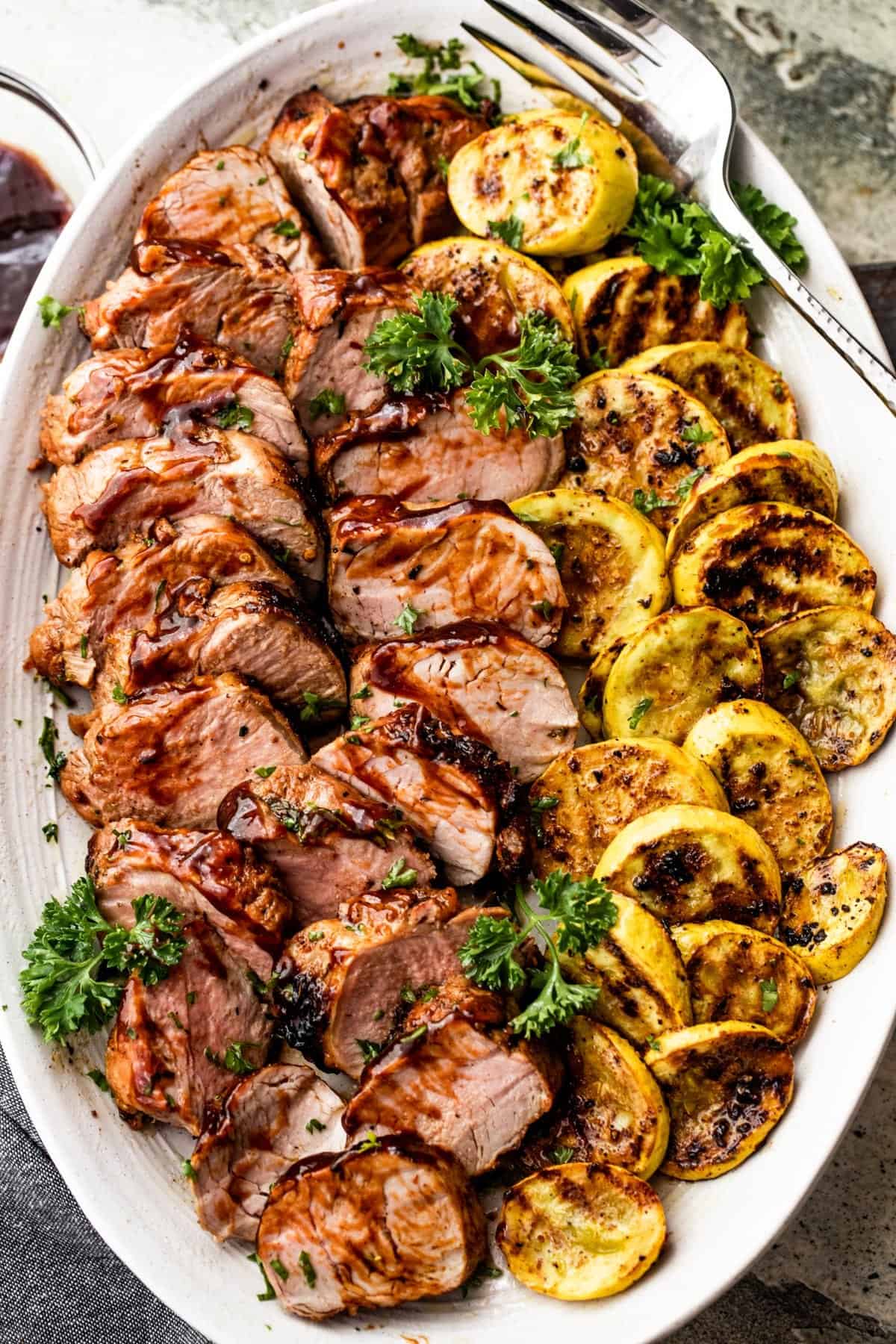 grilled pork tenderloin, sliced, and arranged on an oval platter alongside sliced and grilled yellow squash.