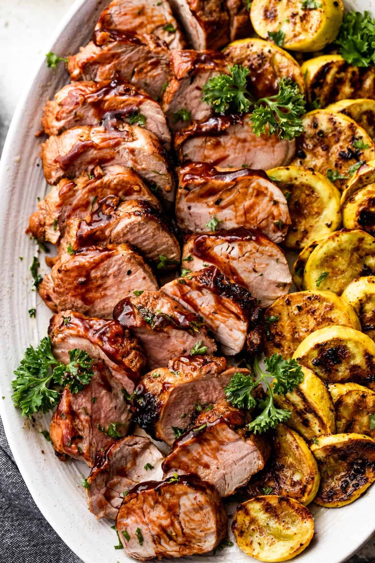 grilled pork tenderloin, sliced, and arranged on an oval platter alongside sliced and grilled yellow squash.