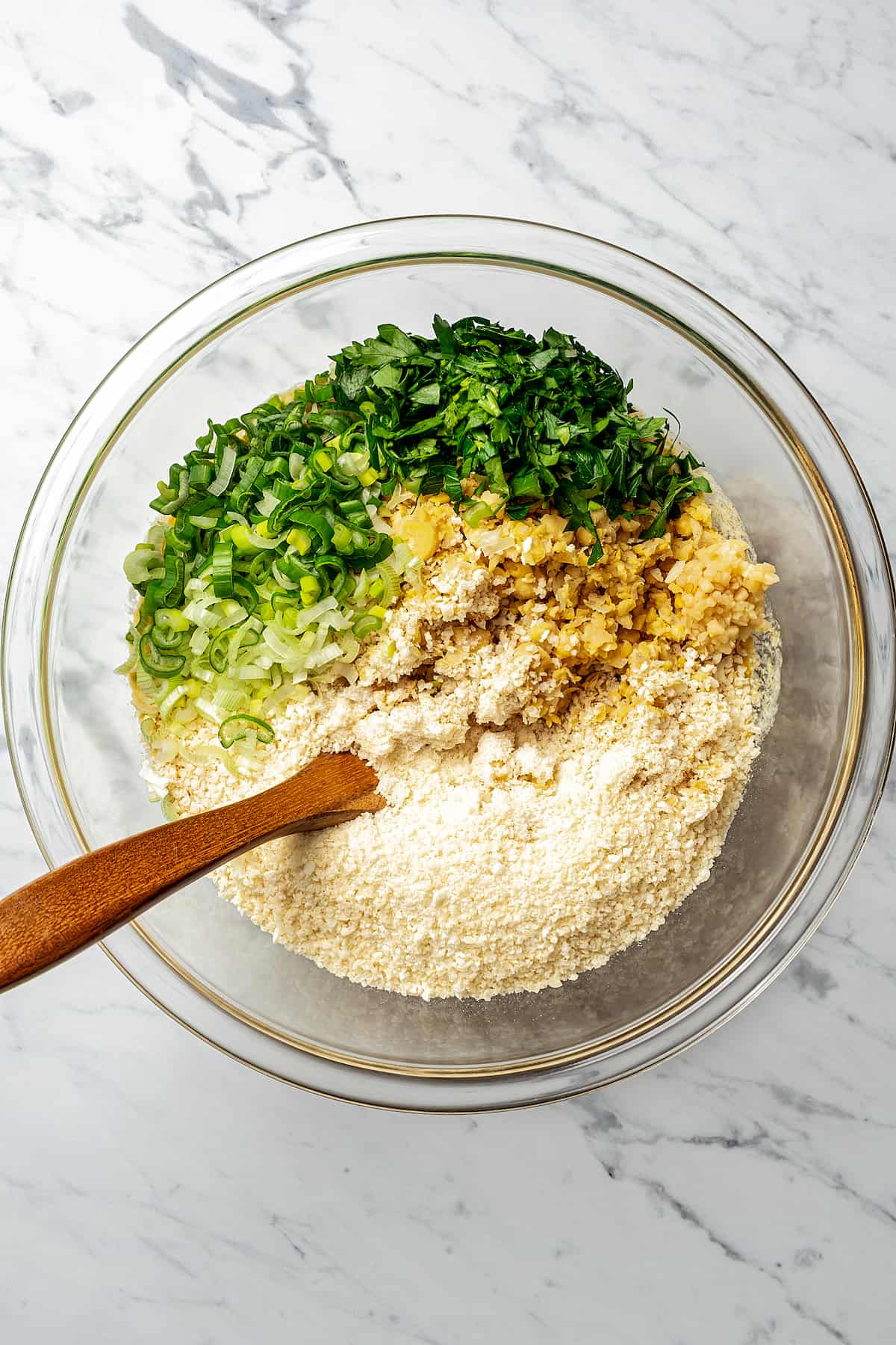 Parsley, breadcrumbs, ground chickpeas, and other ingredients in a glass bowl with a wooden spoon.