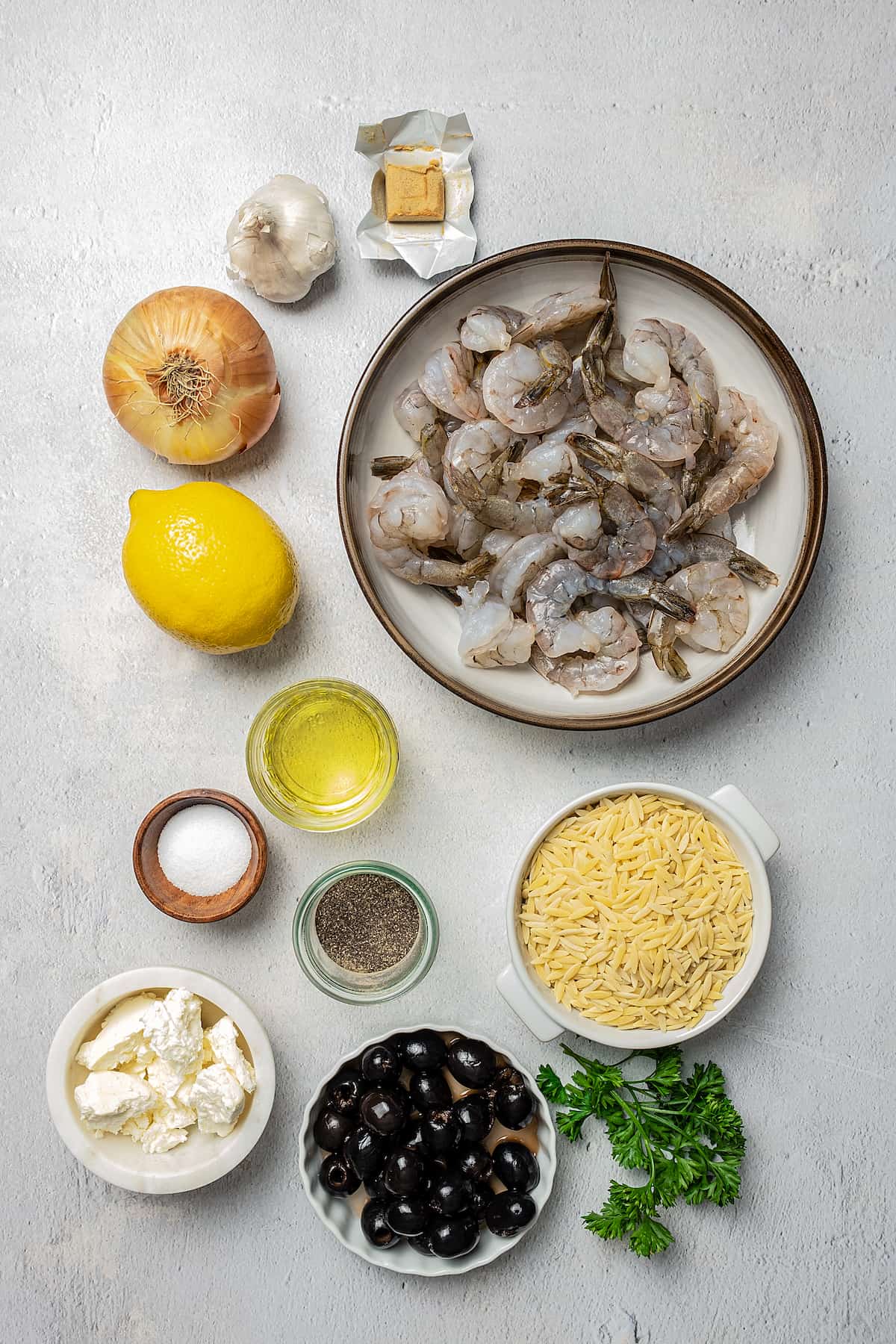 From top left: Seasoning cube, peeled shrimp, garlic and onion, lemon, olive oil, salt, pepper, orzo, feta, black olives, herbs.