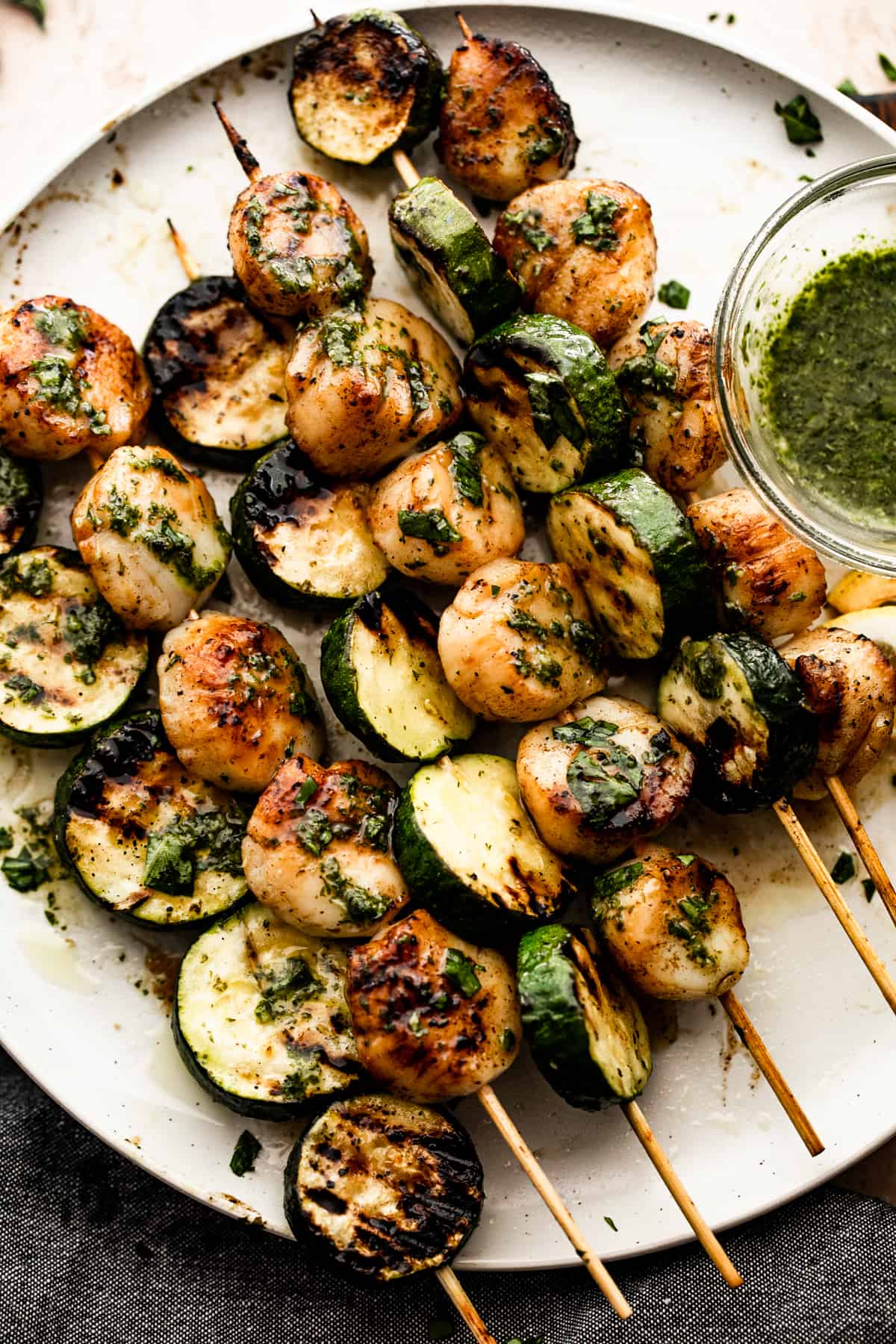 grilled zucchini and grilled scallops threaded onto five skewers and arranged on a white plate with a small bowl of basil and garlic sauce placed next to them.