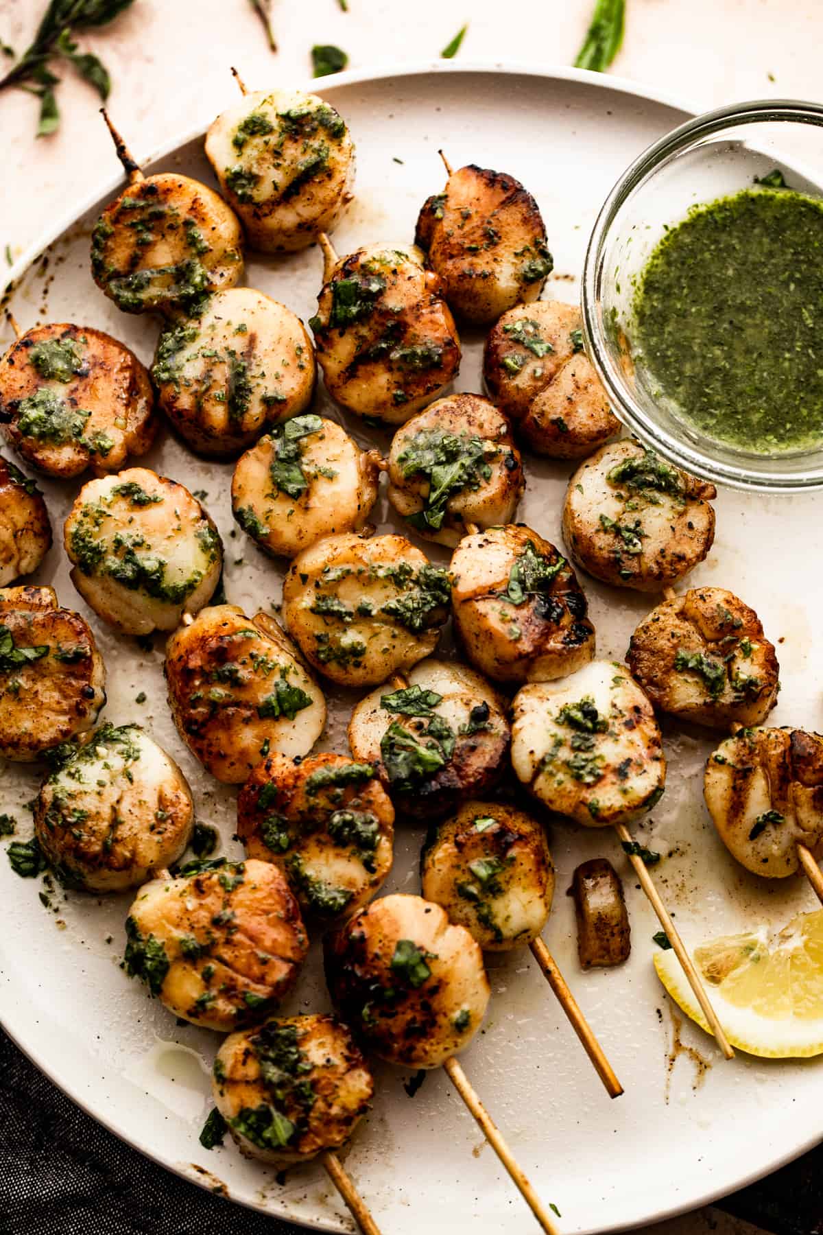grilled zucchini and grilled scallops threaded onto five skewers and arranged on a white plate with a small bowl of basil and garlic sauce placed next to them.