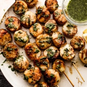 grilled scallops threaded onto five skewers and arranged on a white plate with a small bowl of basil and garlic sauce placed next to them.